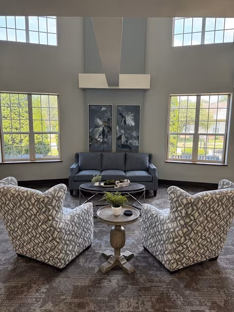A cozy sitting area in a retirement community with a gray sofa against a wall featuring two floral paintings. Two patterned armchairs face the sofa with a small round wooden table between them holding a potted plant and a small dish. Large windows on either side let in natural light and show greenery outside.