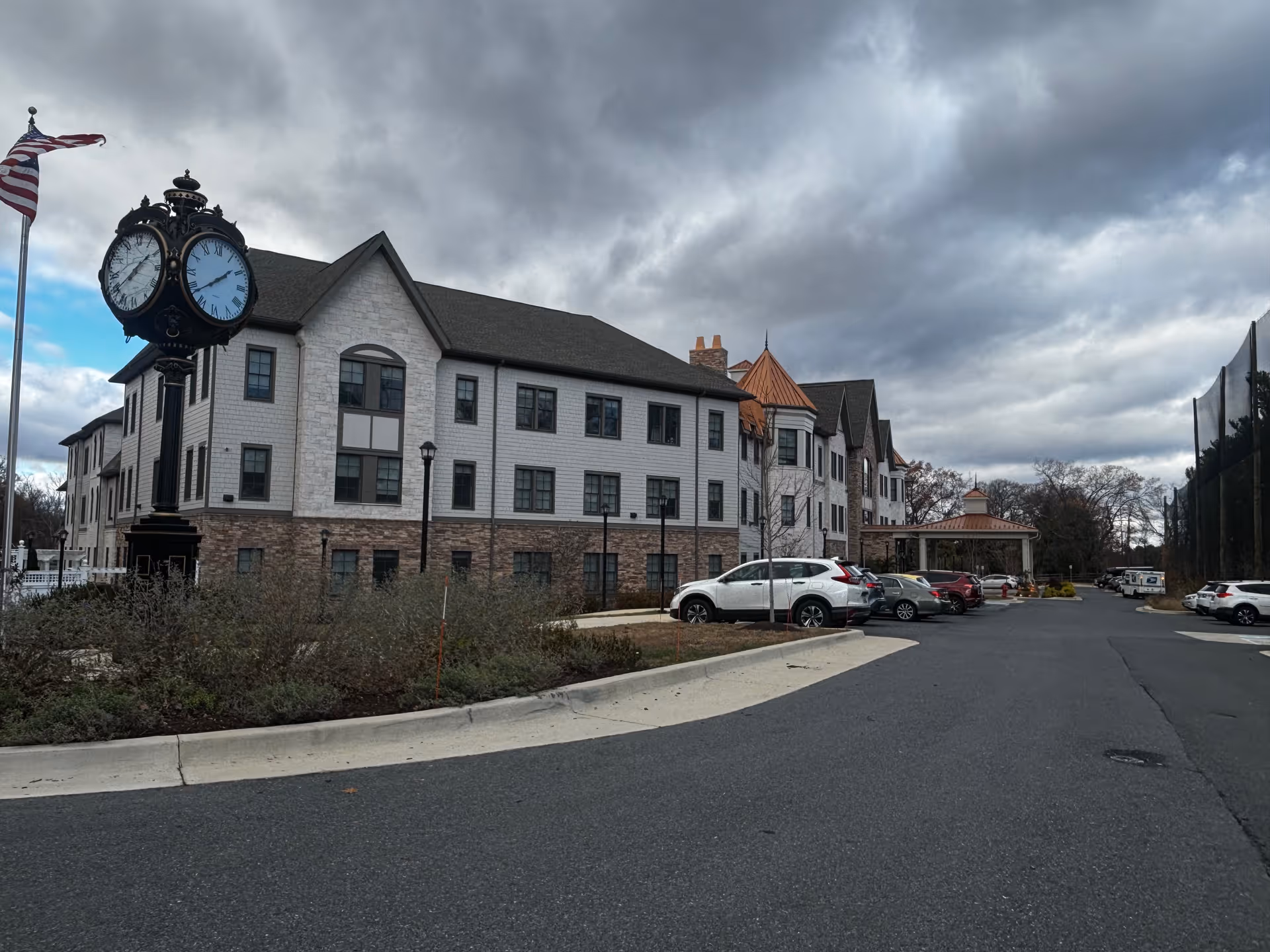 Exterior view of a senior living facility building with a large clock on a pole and an American flag on a flagpole. Several cars are parked along the driveway leading to the building entrance under a cloudy sky.