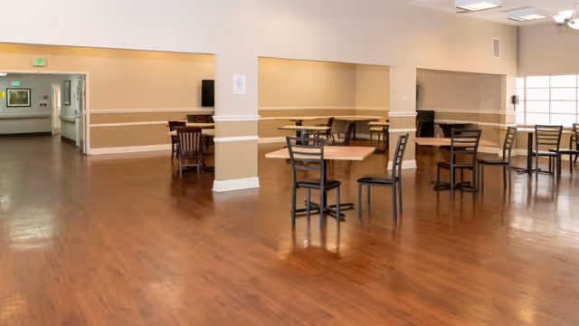 Spacious dining area with wooden floors, several square tables and chairs arranged throughout the room, beige walls with white trim, and a large window allowing natural light to fill the space.