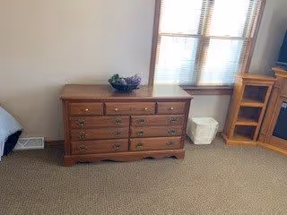 A wooden dresser with multiple drawers placed against a beige wall. On top of the dresser is a decorative bowl with purple flowers. To the right of the dresser is a white wastebasket and a wooden shelving unit. A window with blinds is above the wastebasket, allowing natural light into the room. The floor is carpeted in a neutral color.