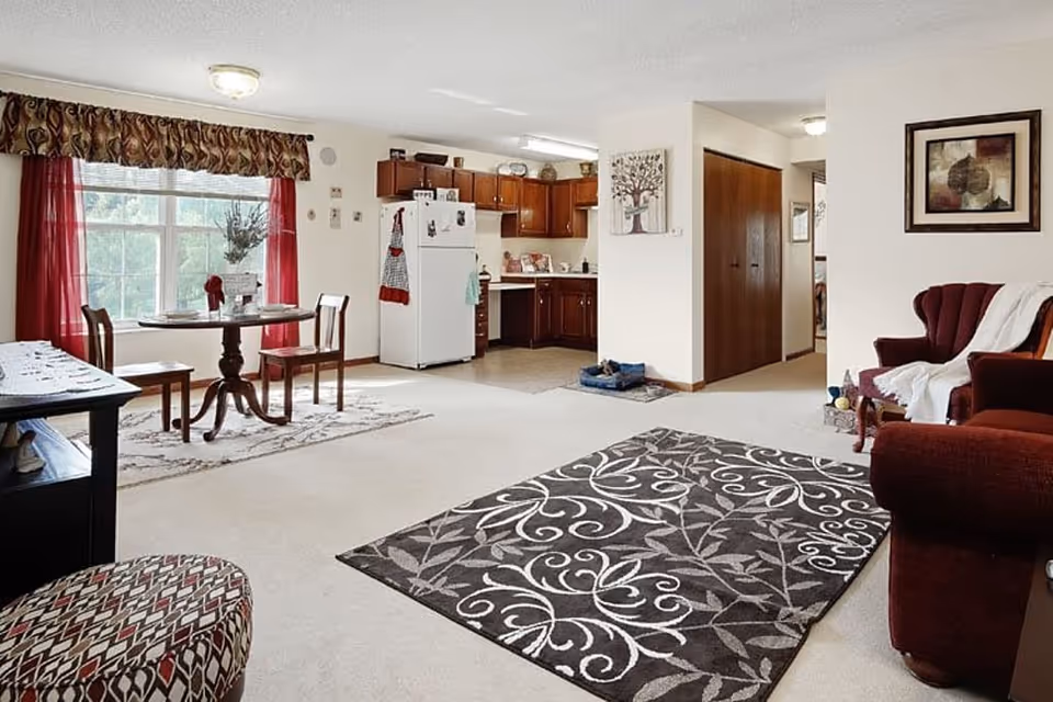 A bright and cozy living area in a senior living facility featuring a patterned area rug on a light carpet, a maroon armchair with a white throw, a small round dining table with two chairs near a large window with red curtains, and a kitchen area with wooden cabinets and a white refrigerator in the background.