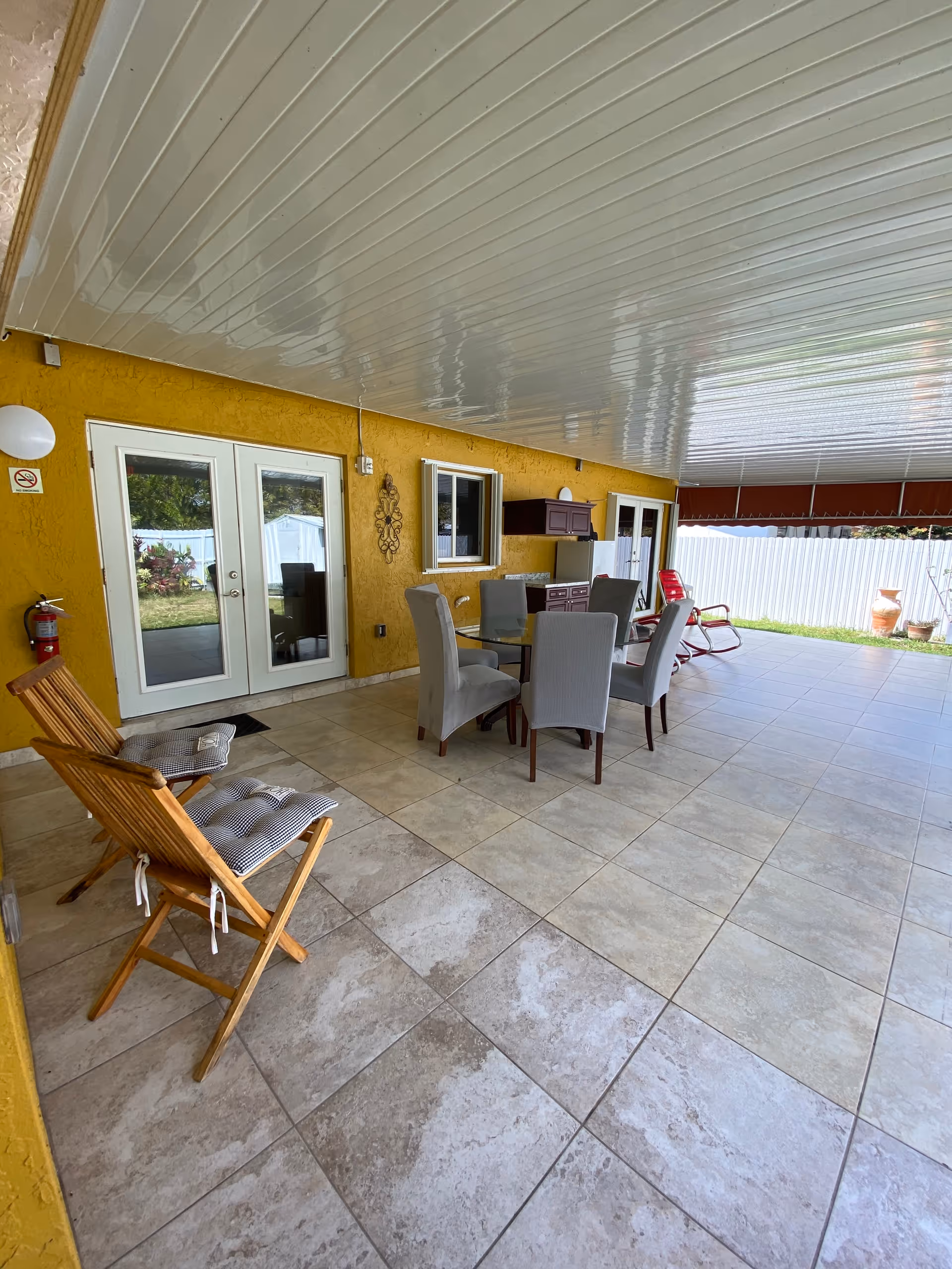 Covered outdoor patio area with tiled floor and yellow walls. There is a glass-top table surrounded by six gray upholstered chairs in the center. Two wooden chairs with cushions are positioned on the left side near a fire extinguisher mounted on the wall. The patio has a white ceiling and is adjacent to a grassy yard enclosed by a white fence. There are double glass doors and a window on the yellow wall, along with a small cabinet mounted on the wall.