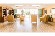 A spacious and bright living room with light wood flooring, beige armchairs arranged around a central area, large windows with green curtains letting in natural light, and wooden furniture including a cabinet and side tables with decorative items.