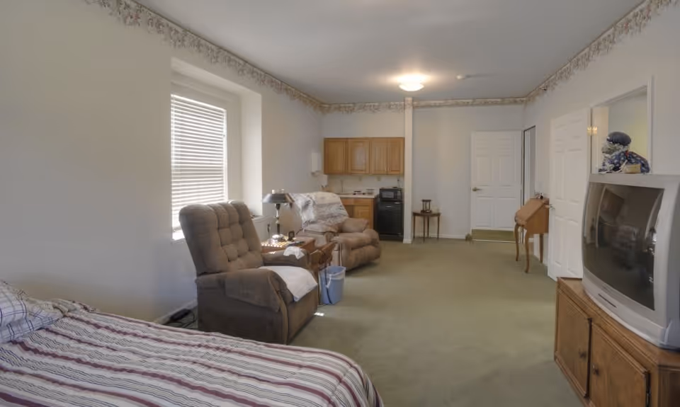 A spacious room with a bed on the left side, two cushioned recliner chairs near a window with blinds, a small wooden table with a lamp between the chairs, a kitchenette with wooden cabinets and a microwave in the background, a TV on a wooden stand on the right, and a small desk near a closed door. The room has light-colored walls with floral border wallpaper near the ceiling and green carpeted floor.