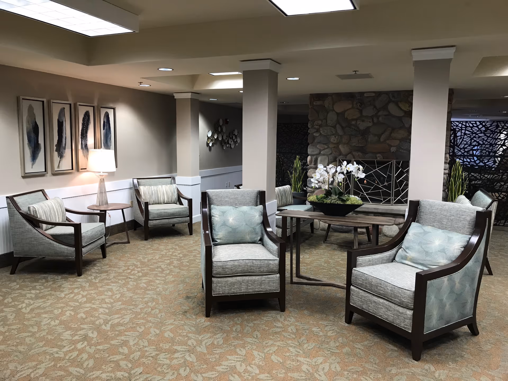 A cozy seating area in a senior living facility with several cushioned armchairs arranged around small tables. The room features a stone fireplace with a decorative metal screen, potted plants, and framed abstract artwork on the walls. The carpet has a leaf pattern, and the lighting is soft and warm.