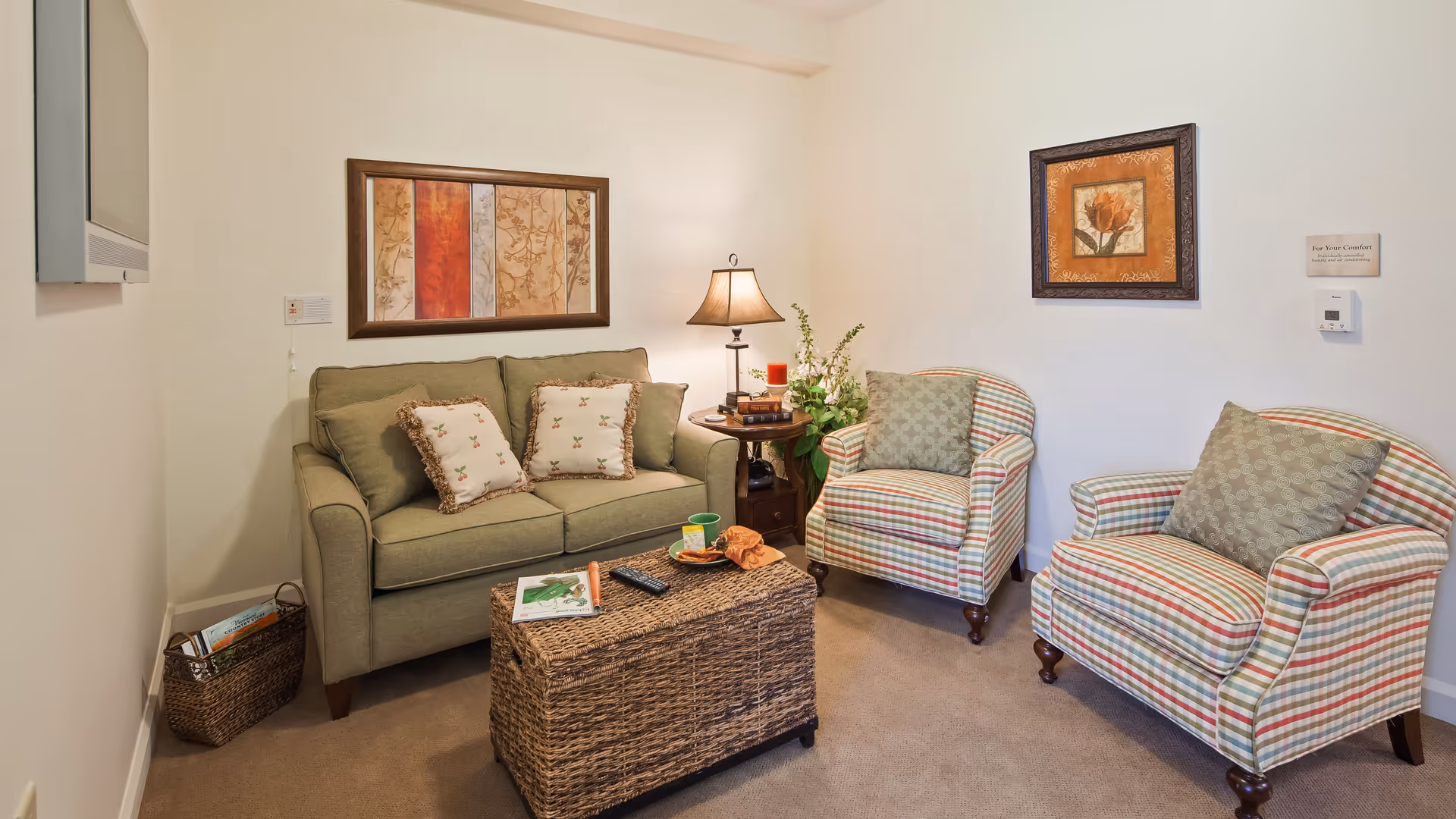 A cozy living room with a green loveseat adorned with two floral pillows, two striped armchairs with green cushions, a wicker coffee table with a magazine, remote control, and a cup on it. A side table with a lamp, books, and a plant is placed between the loveseat and one armchair. The walls are decorated with framed artwork, and a flat-screen TV is mounted on the wall.
