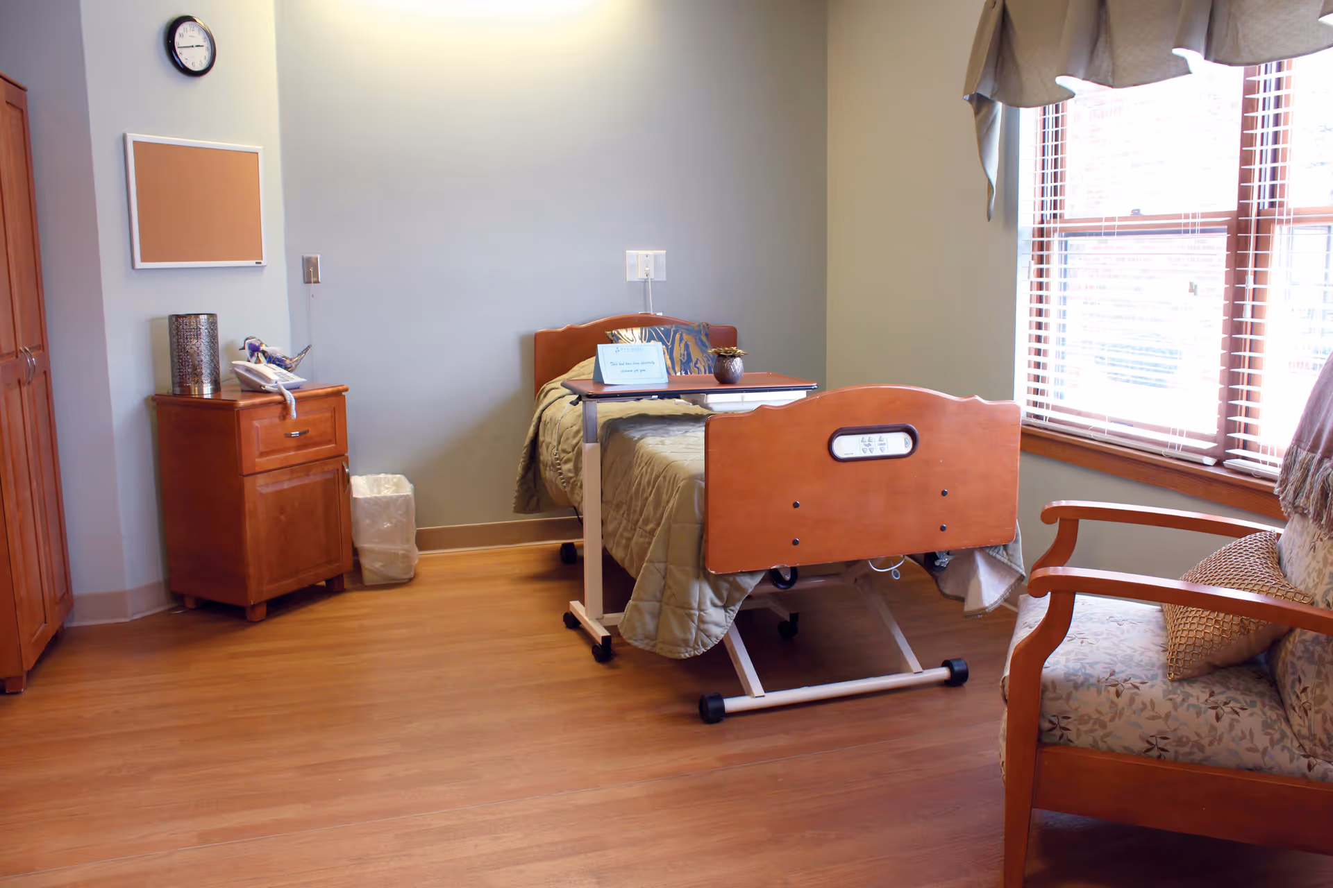 A tidy senior resident bedroom with a hospital-style bed, bedside cabinet, armchair, and large window.