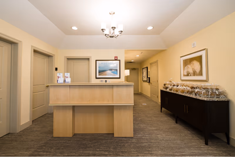 Reception area with a light wood front desk, beige walls, and carpeted floor. There are three closed doors behind the desk, a framed picture of a beach scene on the wall, and a chandelier hanging from the ceiling. To the right, there is a dark wood sideboard with multiple wrapped baked goods and a framed picture of trees above it.