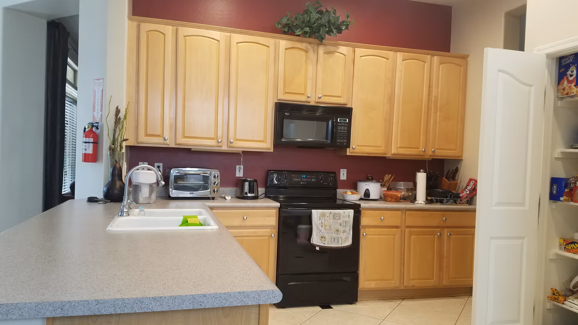 A kitchen with light wood cabinets, a black stove and microwave, a toaster oven, a coffee maker, and a white sink with a faucet on a gray countertop. There is a pantry door open on the right side showing various food items. A fire extinguisher is mounted on the wall to the left, and a decorative plant is placed above the cabinets.