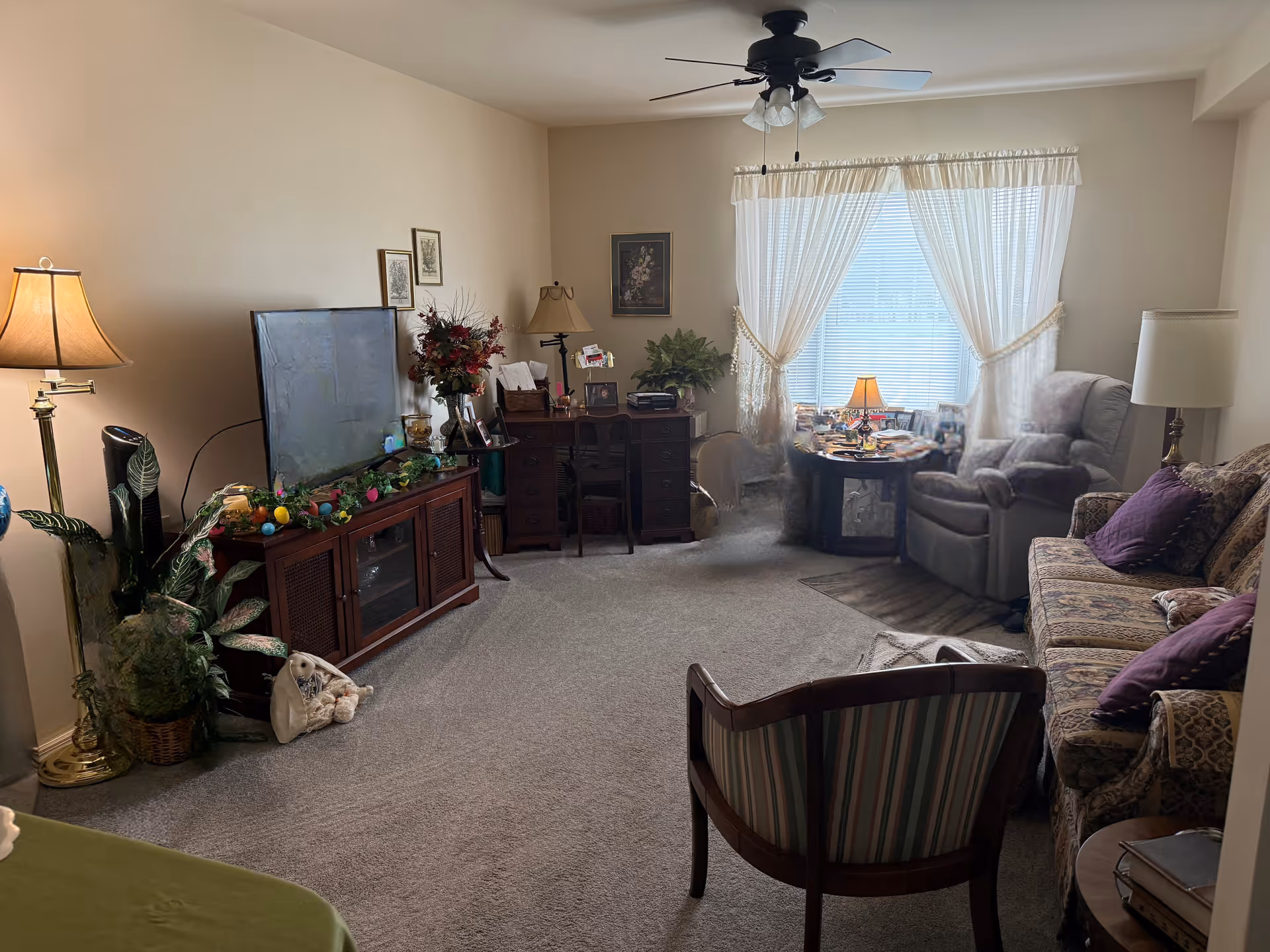 A cozy living room with beige walls and carpeted floor. The room features a large window with sheer white curtains, a ceiling fan with lights, a floral-patterned sofa with purple cushions, a striped armchair, and a recliner chair near a small round table with a lamp. There is a wooden TV stand with a flat-screen TV, decorative plants, and a desk with a chair and various items on it.
