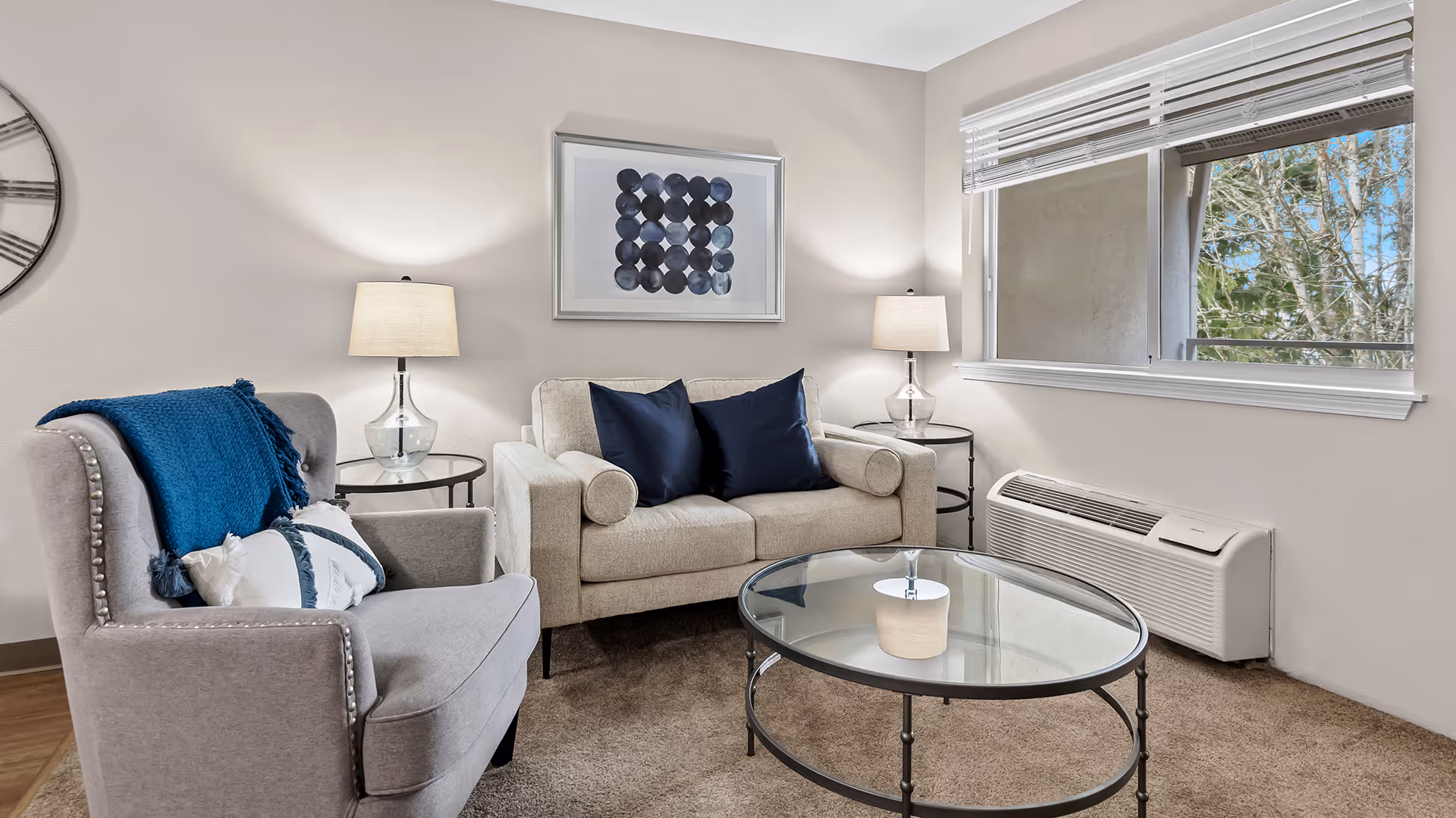 A cozy living room with a beige loveseat adorned with two dark blue pillows, a gray armchair with a blue throw blanket and white pillow, two glass side tables each with a lamp, a round glass coffee table, a large wall clock on the left wall, abstract artwork above the loveseat, and a window with white blinds showing trees outside.