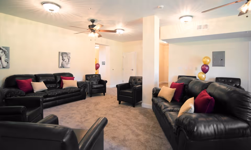 A cozy living room area with black leather sofas and armchairs arranged around a carpeted floor. The sofas have red and beige throw pillows. The walls are light-colored with two black and white portraits hanging. There are ceiling lights and a ceiling fan, and some balloons are visible in the background near a doorway.