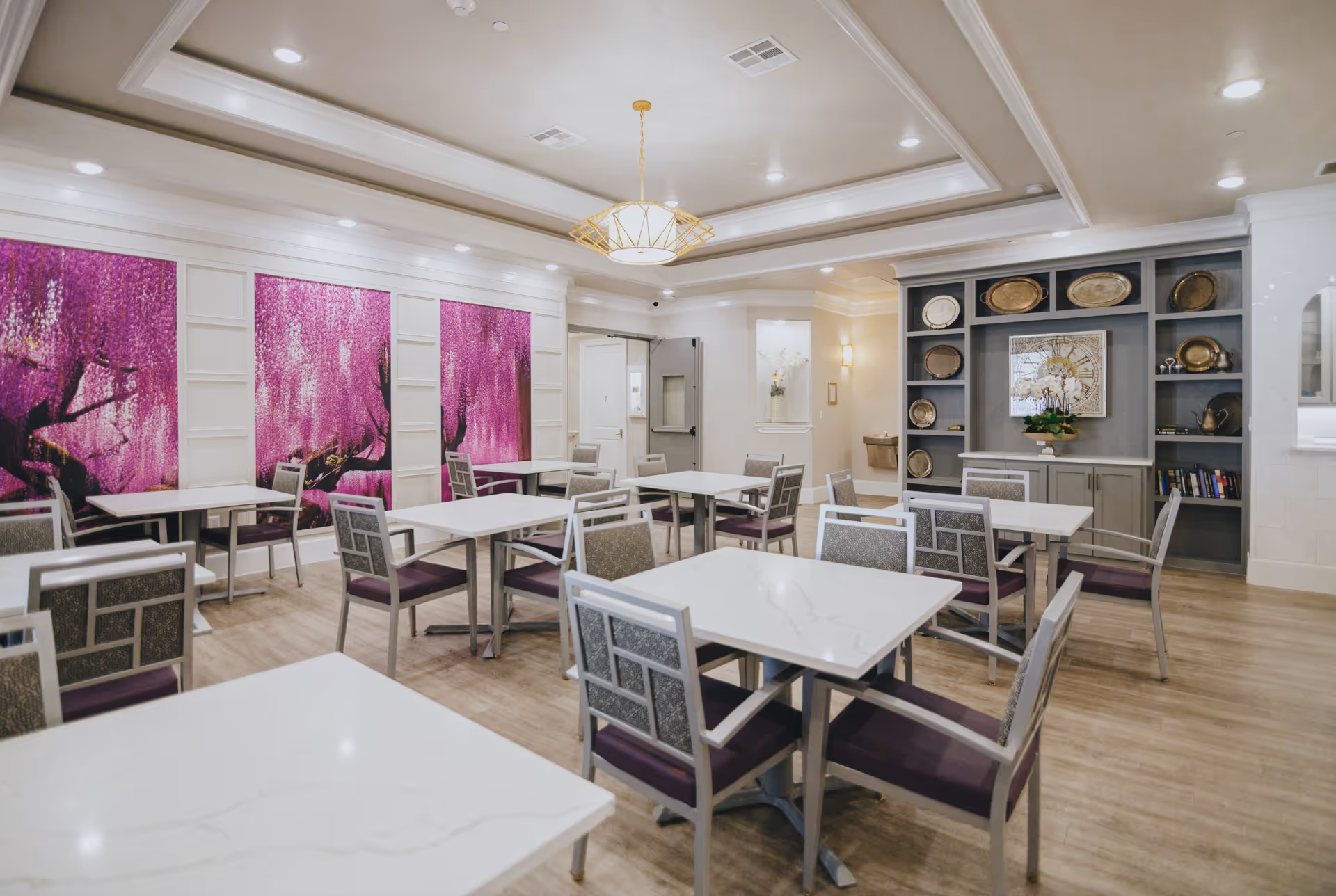 A bright and clean dining room with multiple white marble tables and gray chairs with purple cushions. The room features a decorative ceiling light fixture, a wall with large purple floral artwork, and a built-in shelving unit displaying decorative plates and a clock. The floor is wooden, and the room has a calm and inviting atmosphere.