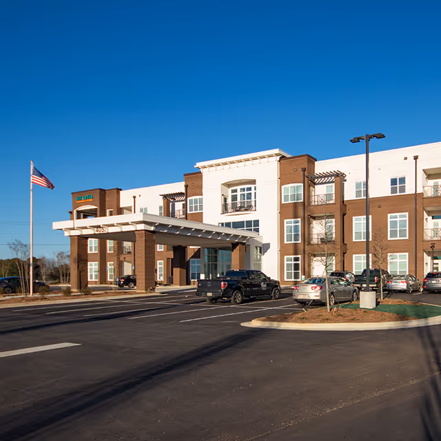 Exterior view of Heartis Suwanee Senior Living facility showing a modern three-story building with brick and white facade, a covered entrance with pillars, several parked cars, a flagpole with an American flag, and clear blue sky.