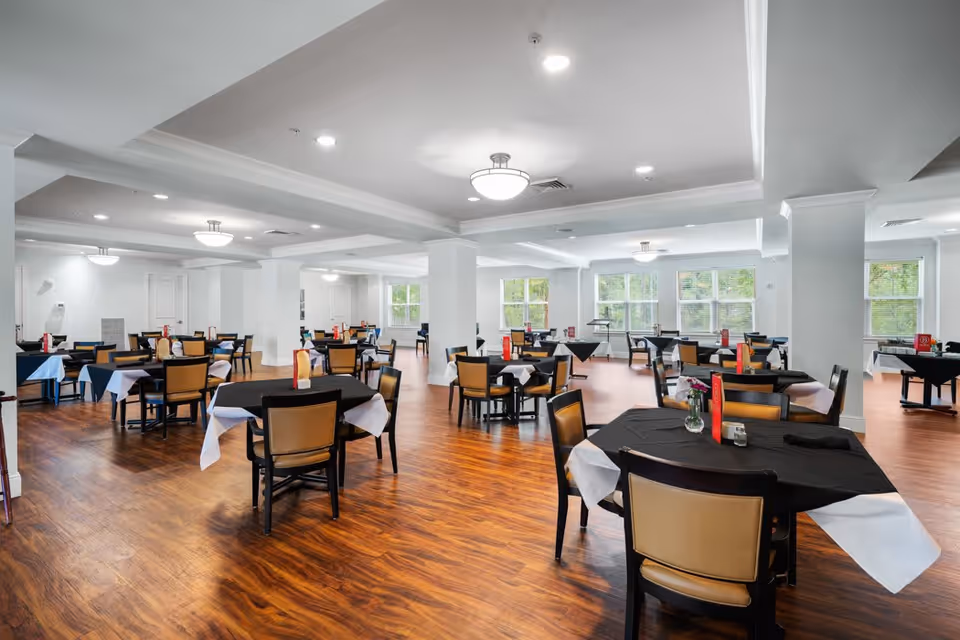 A spacious dining room with multiple round tables covered with black tablecloths and white napkins. Each table is surrounded by tan and black chairs. The room has wooden flooring, white walls, large windows letting in natural light, and ceiling lights providing illumination.