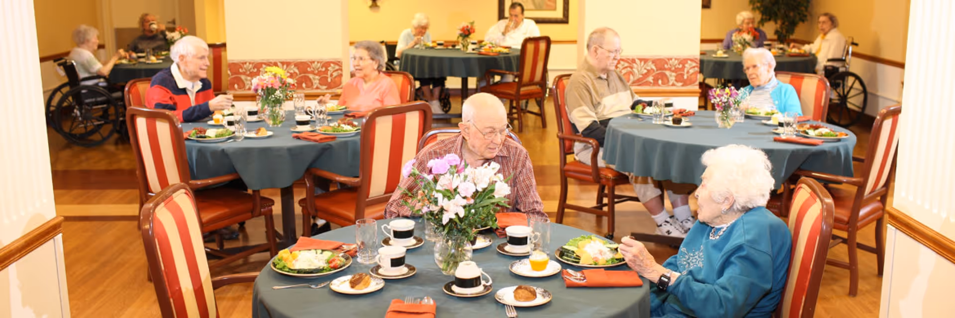 Several elderly residents seated at round dining tables in a well-lit dining room, enjoying meals and engaging in conversation. The tables are covered with dark green tablecloths and decorated with flower arrangements. The room has wooden floors and warm yellow walls with decorative trim.