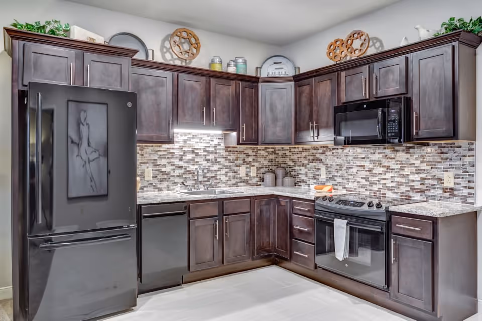 Modern kitchen with dark wooden cabinets, a black refrigerator, dishwasher, stove with oven, and microwave. The backsplash features a mosaic tile design in neutral tones, and there are decorative items on top of the cabinets.