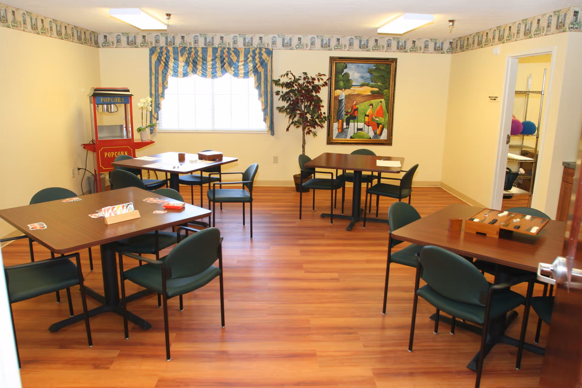 A well-lit activity room with four tables and green chairs arranged around them. On the tables are various game items including playing cards and a backgammon set. A red popcorn machine is positioned near a window with blue and yellow striped curtains. A framed painting and a potted plant are on the wall, and an open door reveals a storage area with shelves and colorful balls.