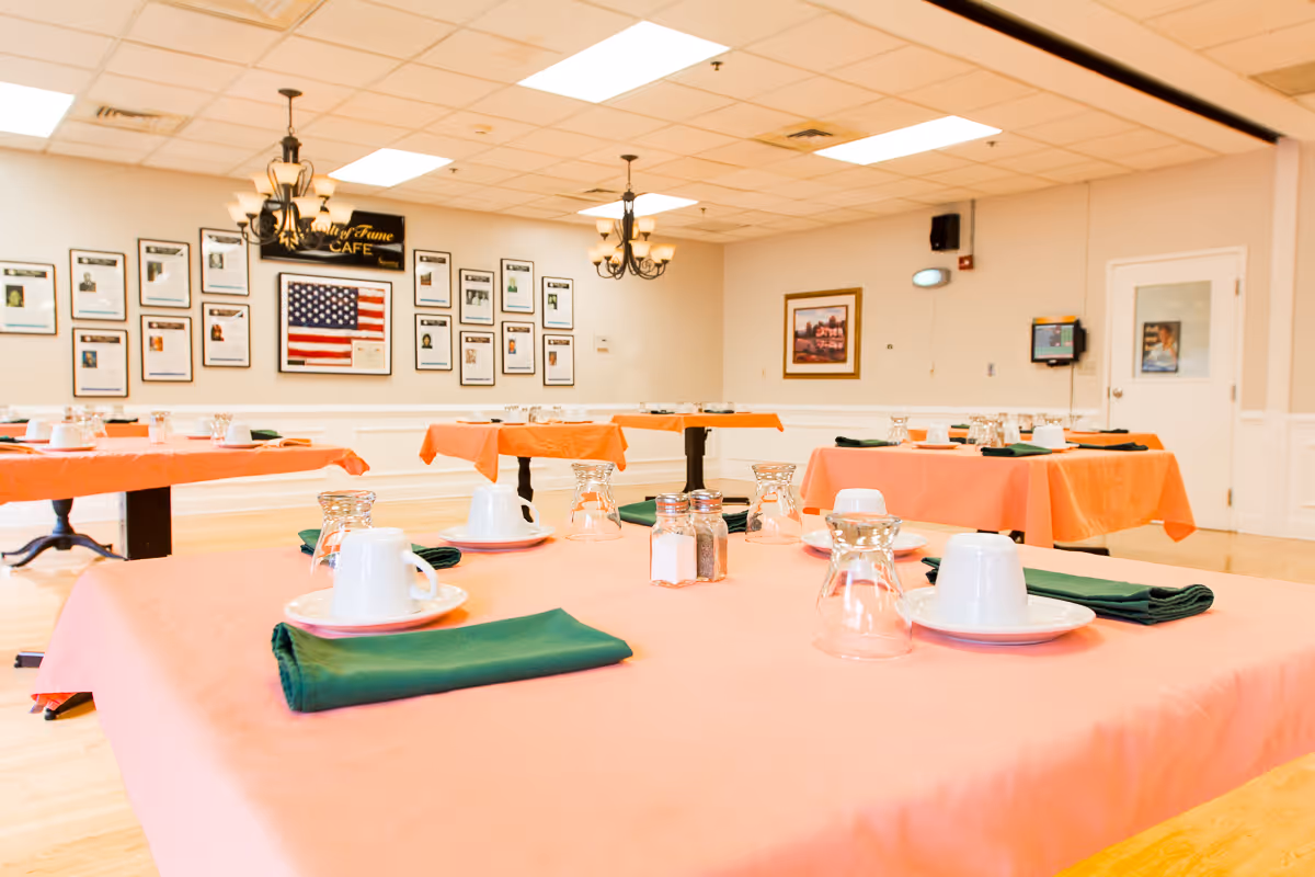 A dining room with several tables covered in orange tablecloths, each set with white cups, saucers, upside-down glasses, green napkins, and salt and pepper shakers. The walls have framed photos and an American flag, with chandeliers hanging from the ceiling.