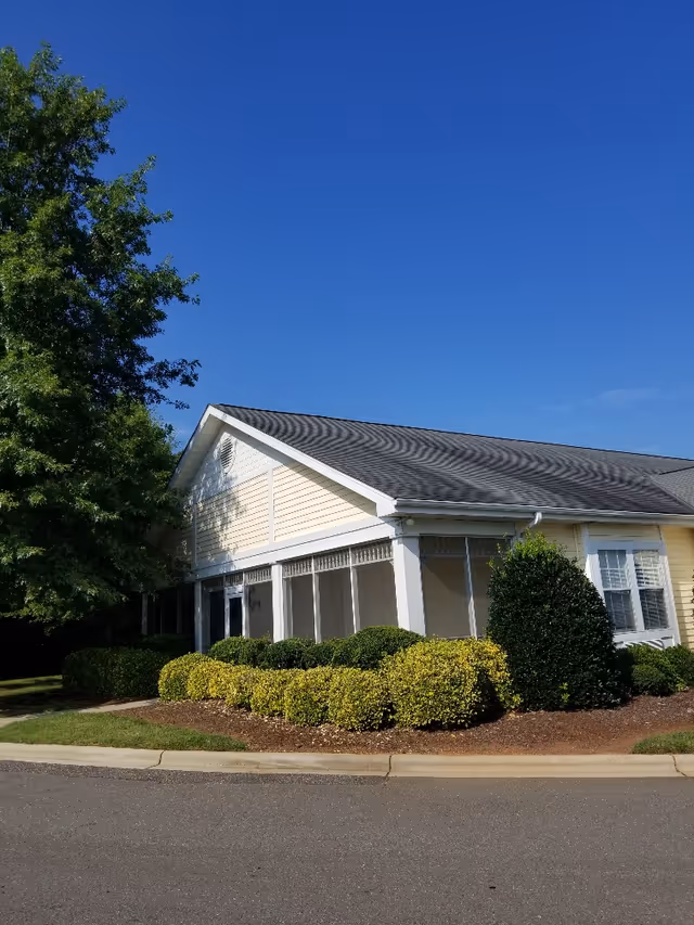 Exterior view of a single-story building with a light yellow facade, white trim, and a dark shingled roof. The building is surrounded by neatly trimmed bushes and a tree on the left side. The sky is clear and blue.