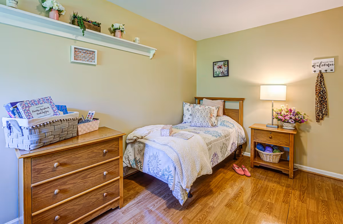 A cozy bedroom with a single bed adorned with floral-patterned bedding and multiple pillows. Next to the bed is a wooden nightstand with a lamp, a basket of yarn, and a vase of flowers. On the left side, there is a wooden dresser with baskets and books on top. The walls are decorated with small framed signs and a shelf holding plants and decor items. The floor is wooden, and a pair of pink slippers is placed beside the bed.