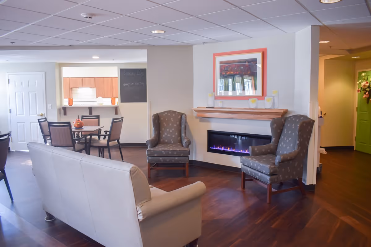 A cozy living room area in a senior living facility featuring two patterned armchairs and a beige sofa arranged around a modern electric fireplace with a wooden mantel. Above the mantel is a framed artwork. In the background, there is a dining area with a table and chairs, and a kitchen pass-through window. The room has wood flooring and a ceiling with recessed lighting.