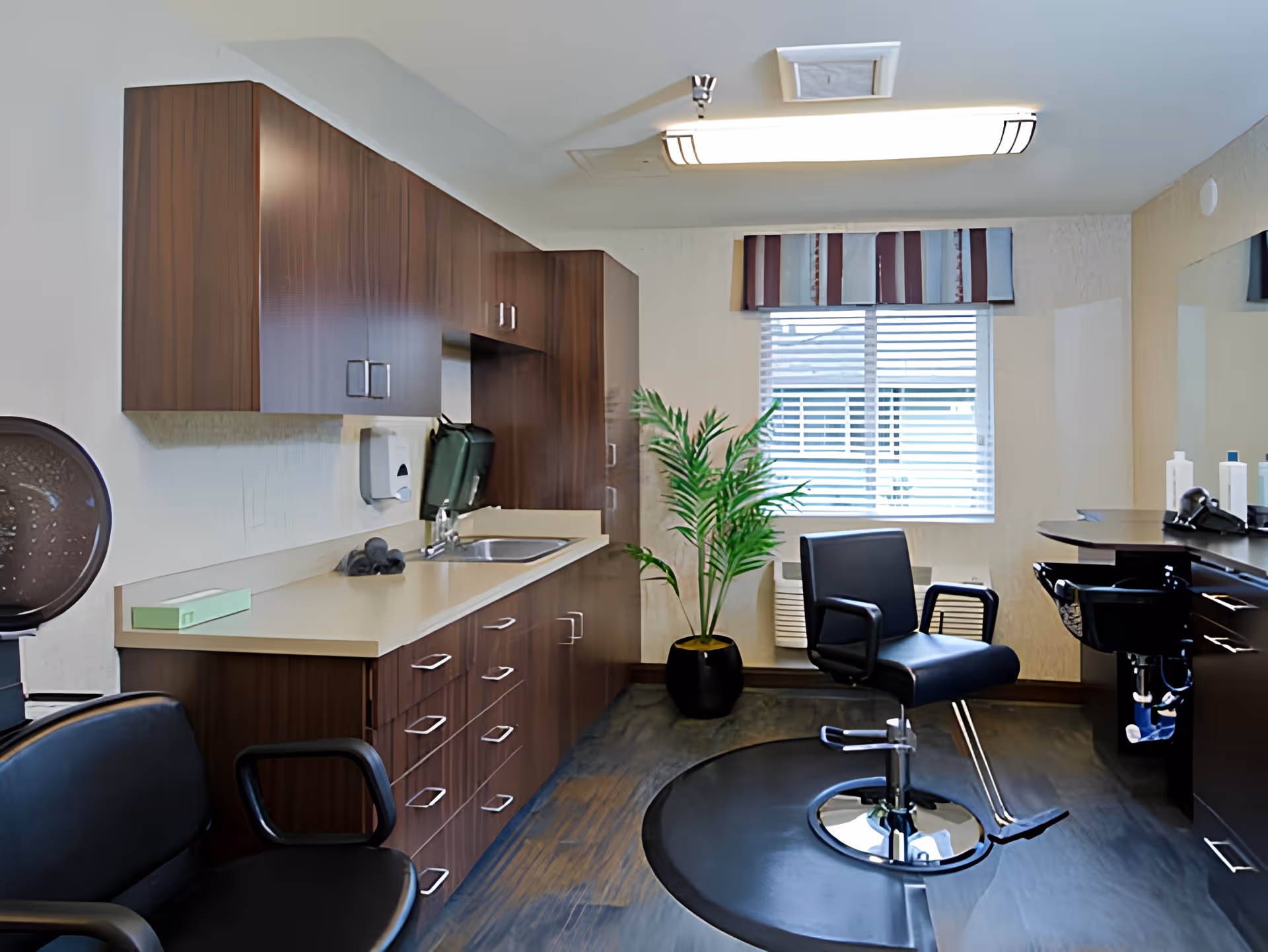 Interior of a salon room with a black styling chair on a circular mat, a black waiting chair, wooden cabinets with a countertop and sink, a potted plant near a window with blinds and a striped valance, and salon equipment on a counter with a large mirror.