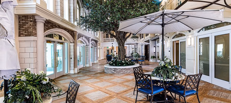 Indoor courtyard area with a large tree in the center surrounded by a circular planter. Several tables with blue cushioned chairs and large white umbrellas are arranged around the space. The area is enclosed by walls with large windows and doors, featuring decorative columns and brick accents.