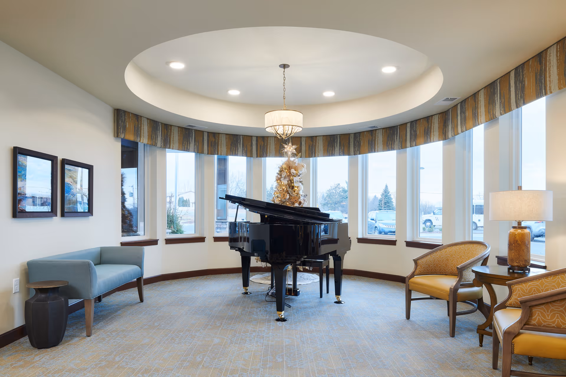 A bright, circular room with large windows and a recessed ceiling light fixture. In the center is a black grand piano decorated with a small Christmas tree on top. The room has a blue cushioned bench on the left, two yellow upholstered chairs on the right, a small side table with a lamp, and framed pictures on the wall.