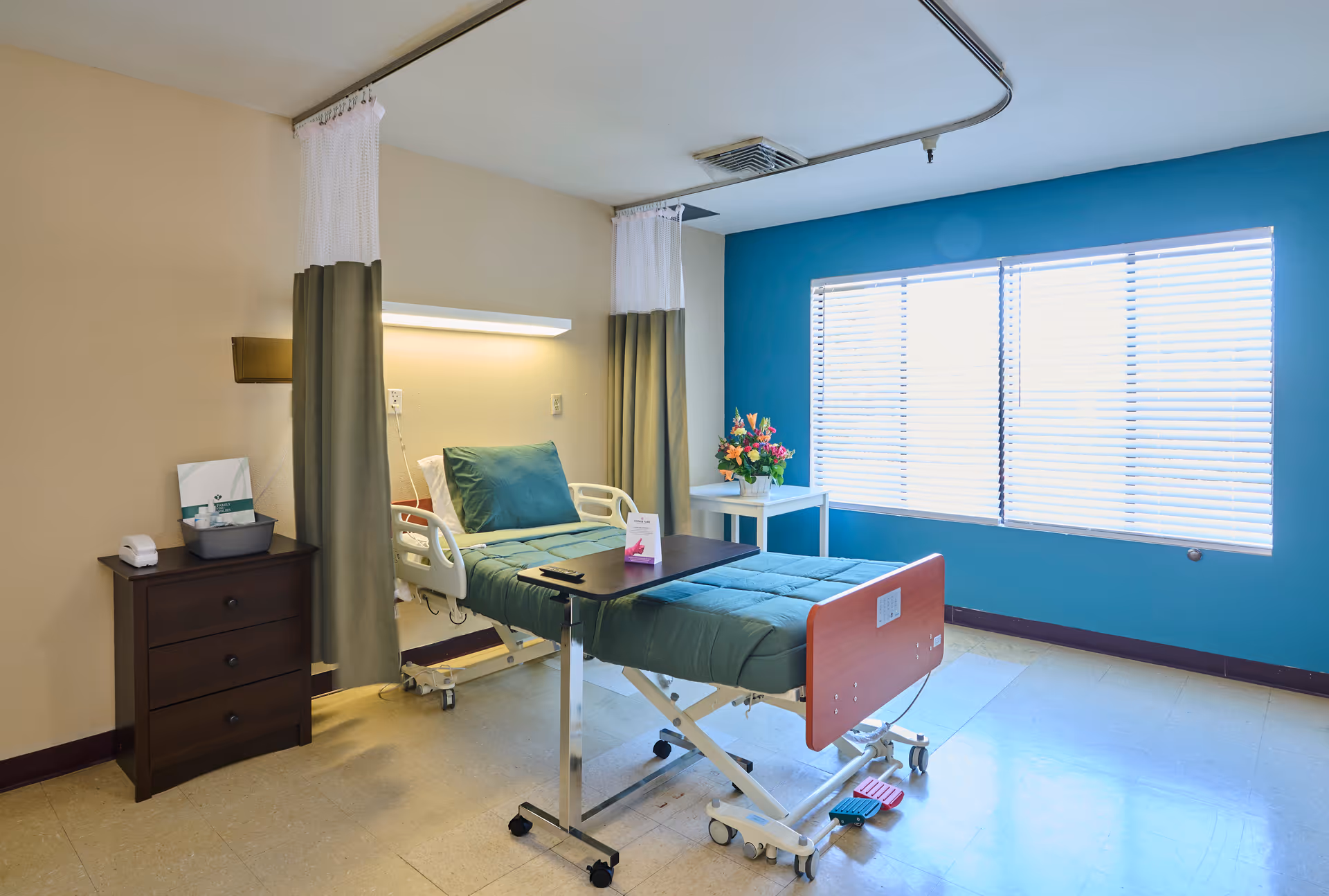 A bright nursing facility room with a hospital bed covered in green bedding, a small bedside table with a tissue box and other items, a white table with a colorful flower arrangement near a large window with blinds, and privacy curtains partially drawn around the bed.