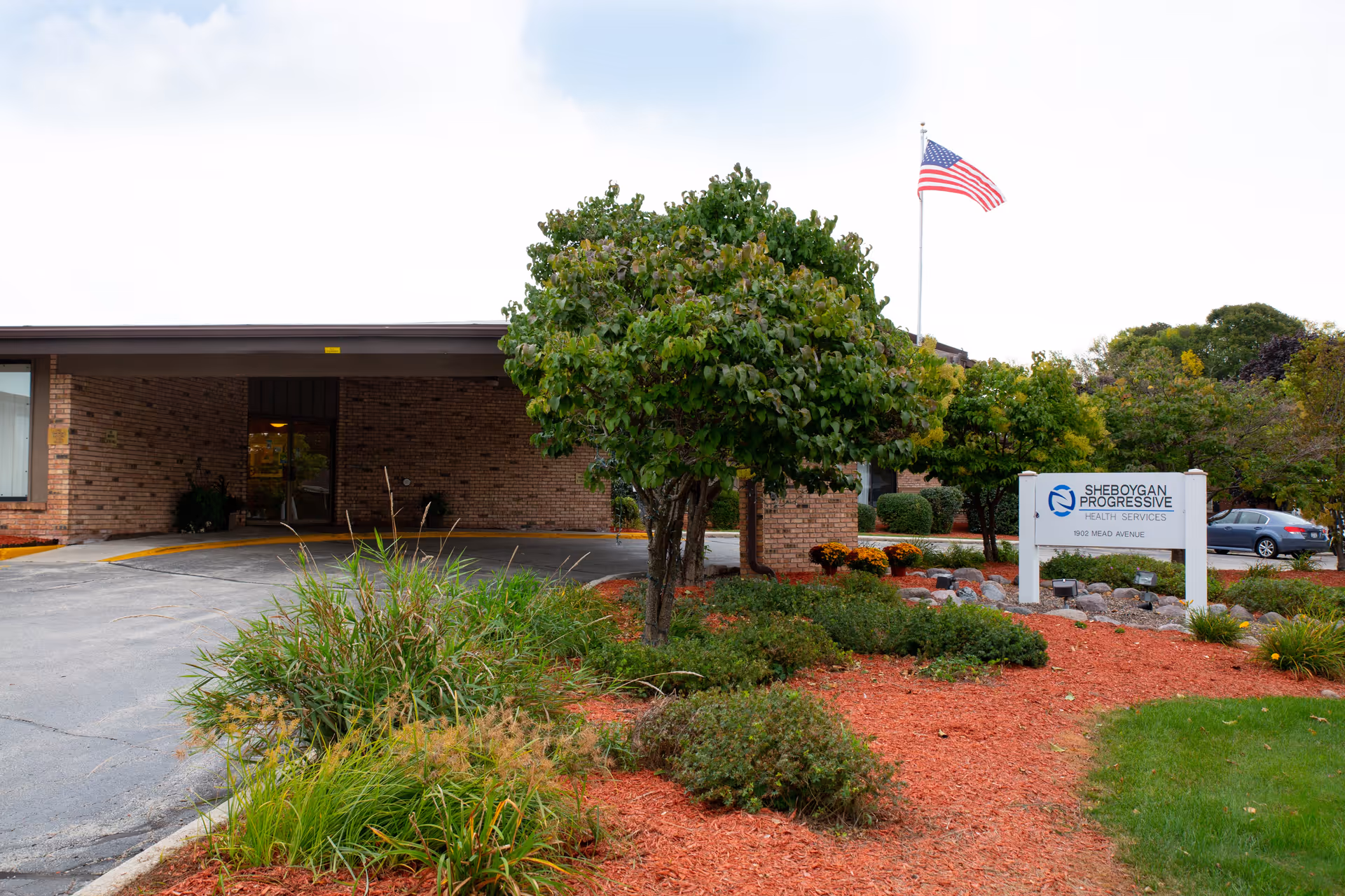 Exterior view of Sheboygan Progressive Health Services building with a brick facade, a covered entrance, landscaped garden with bushes and trees, an American flag on a flagpole, and a sign displaying the facility name and address.