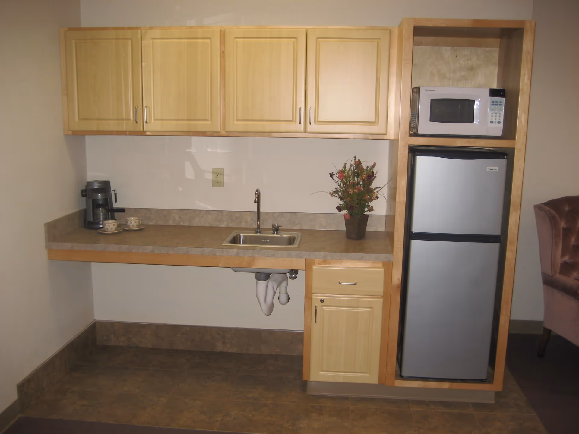 A small kitchenette area with light wood cabinets, a countertop with a sink, a coffee maker with two cups, a flower vase, a microwave, and a stainless steel refrigerator. Part of a pink upholstered chair is visible on the right side.