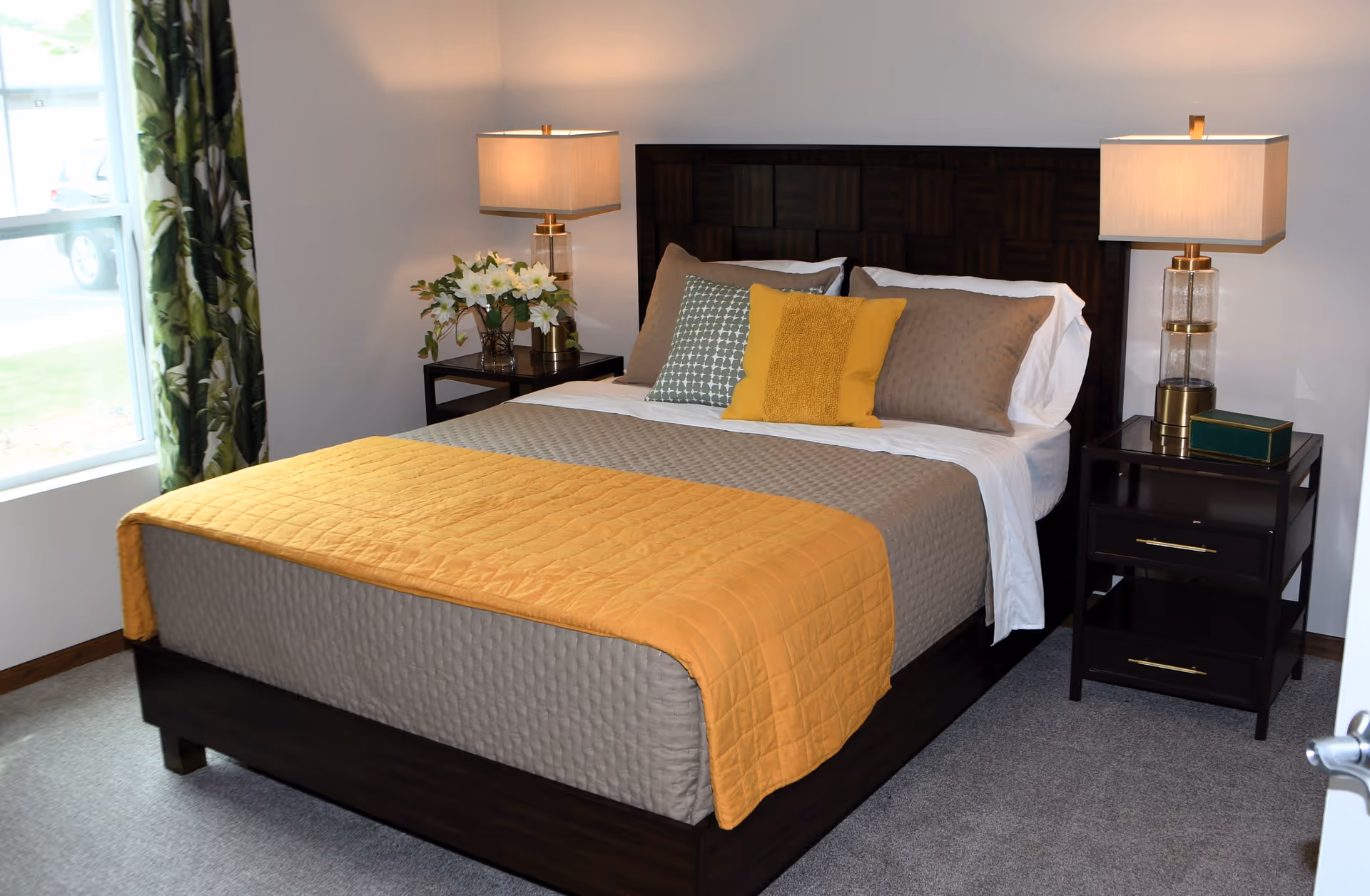 A neatly made bed with a dark wooden headboard, gray quilted bedding, and a mustard yellow quilted throw blanket. The bed has three decorative pillows in gray, green, and yellow. On each side of the bed are dark wooden nightstands with modern table lamps and decorative items. A window with green patterned curtains is visible on the left side of the room.