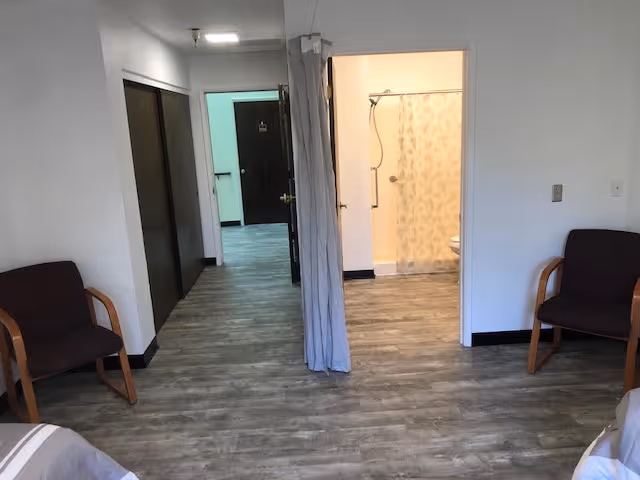 Assisted-living bedroom with wood-look floors, two chairs, a hallway and an open doorway revealing a bathroom with a shower curtain.
