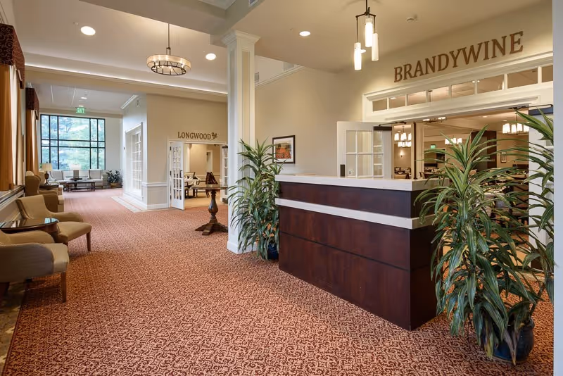 Interior view of a senior living facility hallway with a reception desk on the right labeled 'BRANDYWINE' and an open doorway labeled 'LONGWOOD' in the background. The area is carpeted with a patterned design, has beige walls, several potted plants, and seating areas with chairs along the left side near large windows.