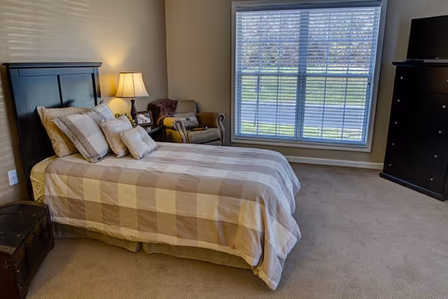 A bedroom with a bed covered in beige and white checkered bedding, a bedside table with a lamp, a cushioned armchair, a large window with blinds, and a tall dark dresser with a TV on top.