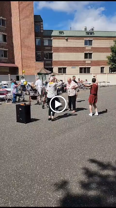 A group of elderly people and caregivers are gathered outside in a paved courtyard area next to a multi-story brick building. Some individuals are dancing while others are seated at tables under umbrellas. The sky is clear and blue.
