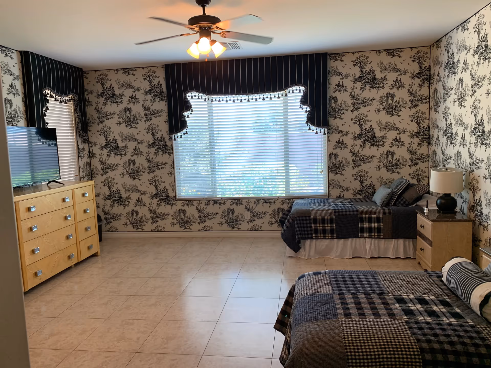 A bedroom with two beds, patterned wallpaper, a dresser, window valances, and a ceiling fan.