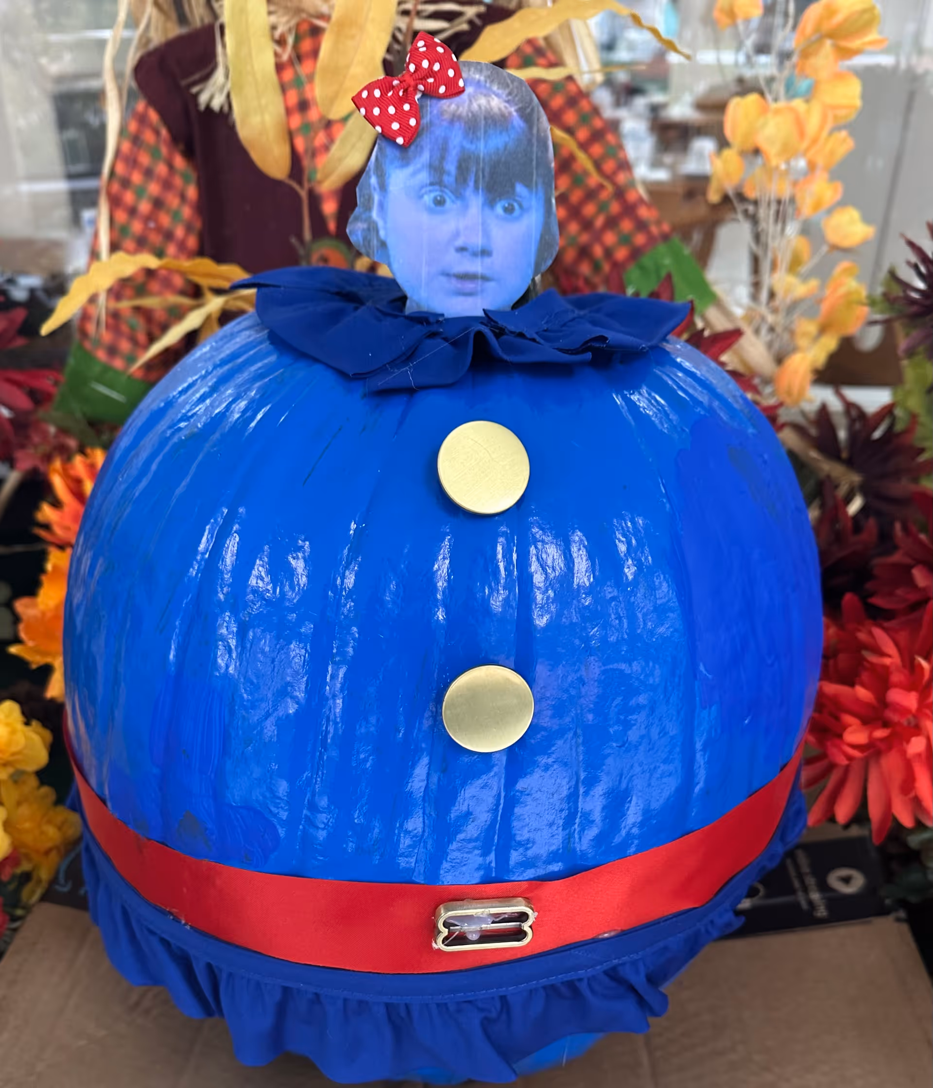 A blue painted pumpkin decorated with gold buttons, a red ribbon belt, ruffled fabric trim, and a cutout photo of a face topped with a red bow, displayed among artificial flowers.