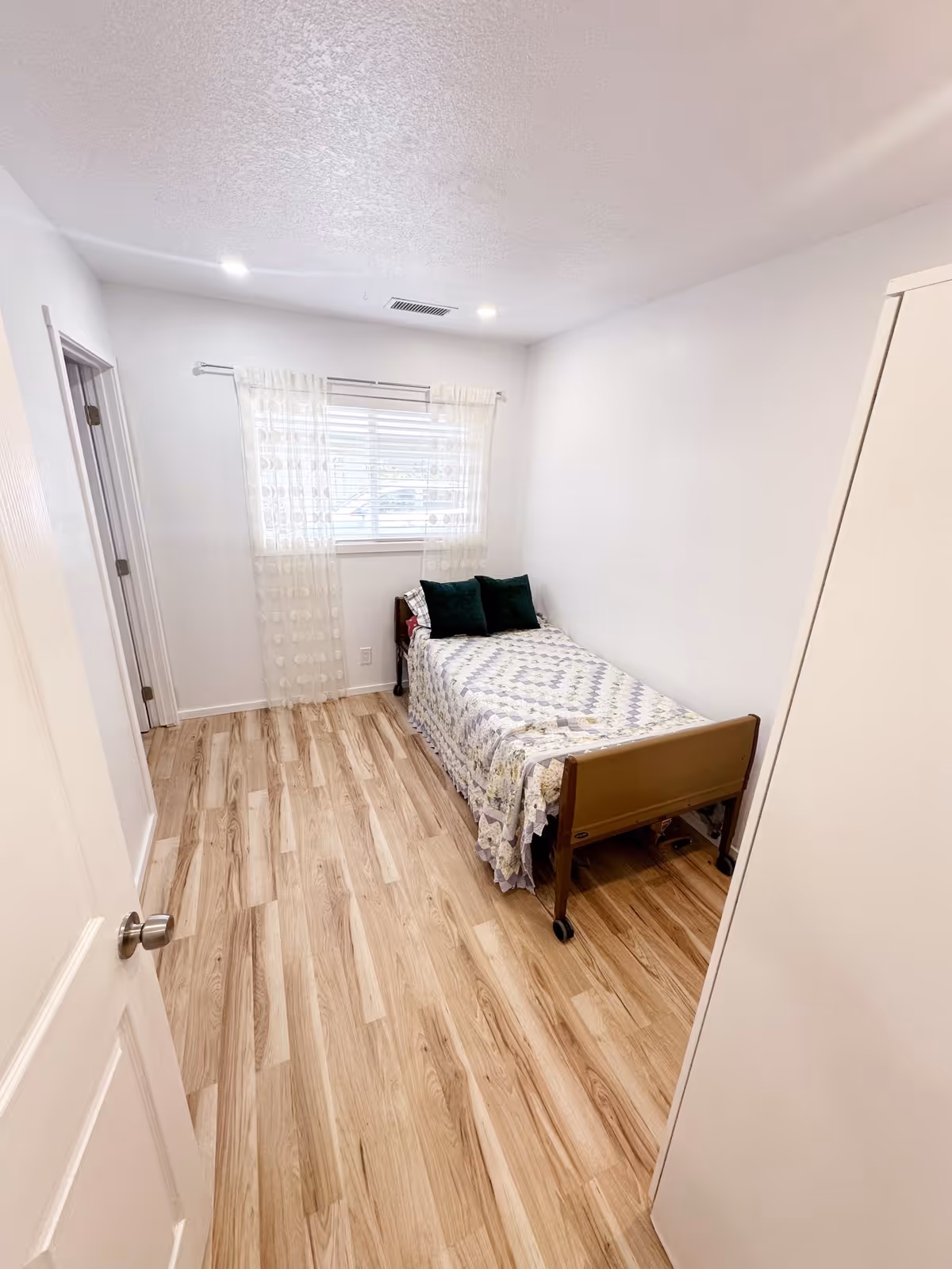 Small bright bedroom with a single bed against the right wall, a sheer-curtained window, and light wood flooring.