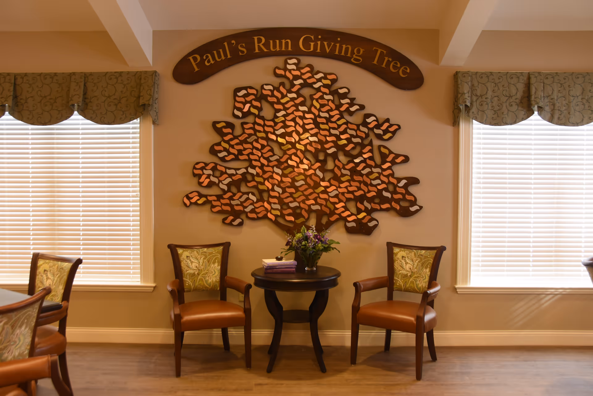 A cozy seating area with two wooden chairs featuring patterned cushions and a small round table between them holding a vase of flowers and two books. Behind the table is a decorative wall piece shaped like a tree with many small plaques, under a wooden sign that reads 'Paul's Run Giving Tree'. Two windows with blinds and valances flank the seating area.
