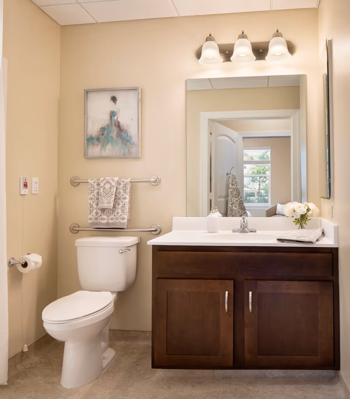 A clean and well-lit bathroom featuring a white toilet with a patterned towel hanging above it on a double towel bar. To the right is a dark wood vanity with a white countertop, a sink, a soap dispenser, a folded towel, and a small vase with white flowers. Above the vanity is a large mirror reflecting a doorway and a window with greenery outside. A three-light fixture with frosted glass shades is mounted above the mirror. A framed artwork of a woman in a blue dress hangs on the wall above the towel bar.