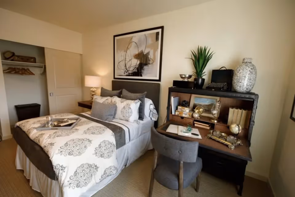 A cozy bedroom featuring a neatly made bed with patterned bedding and multiple pillows, a bedside table with a lamp, an open closet with hangers and a suitcase, and a dark wooden desk with a chair. The desk is decorated with books, a plant, a typewriter, and various decorative items. A large abstract painting hangs above the bed.