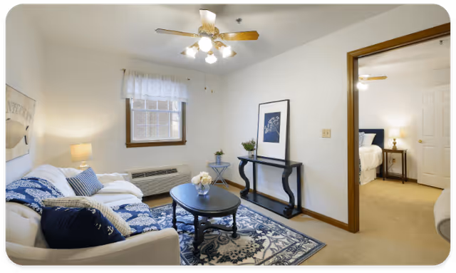 A cozy living room with a white couch adorned with blue and white patterned cushions and a matching throw. A black coffee table with a flower vase sits on a blue and white patterned rug. There is a small black console table with a framed picture and plants against the wall. A window with white curtains and a ceiling fan with lights are visible. An open doorway leads to a bedroom with a bed, nightstand, and lamp.