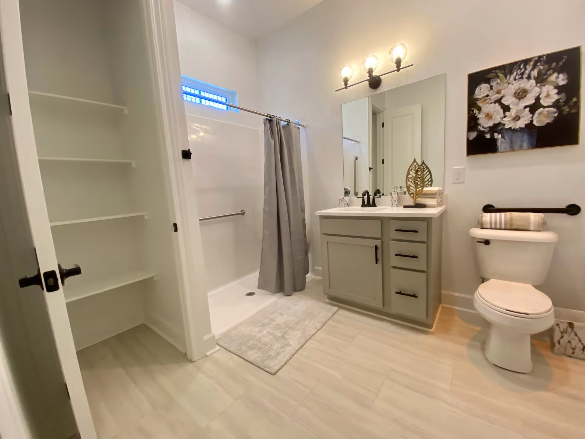 A clean and modern bathroom featuring a walk-in shower with a gray curtain, a vanity with a large mirror and three light bulbs above it, a toilet with a towel on the tank, and a wall-mounted floral painting. There is also an open shelving unit on the left side and light-colored tile flooring.