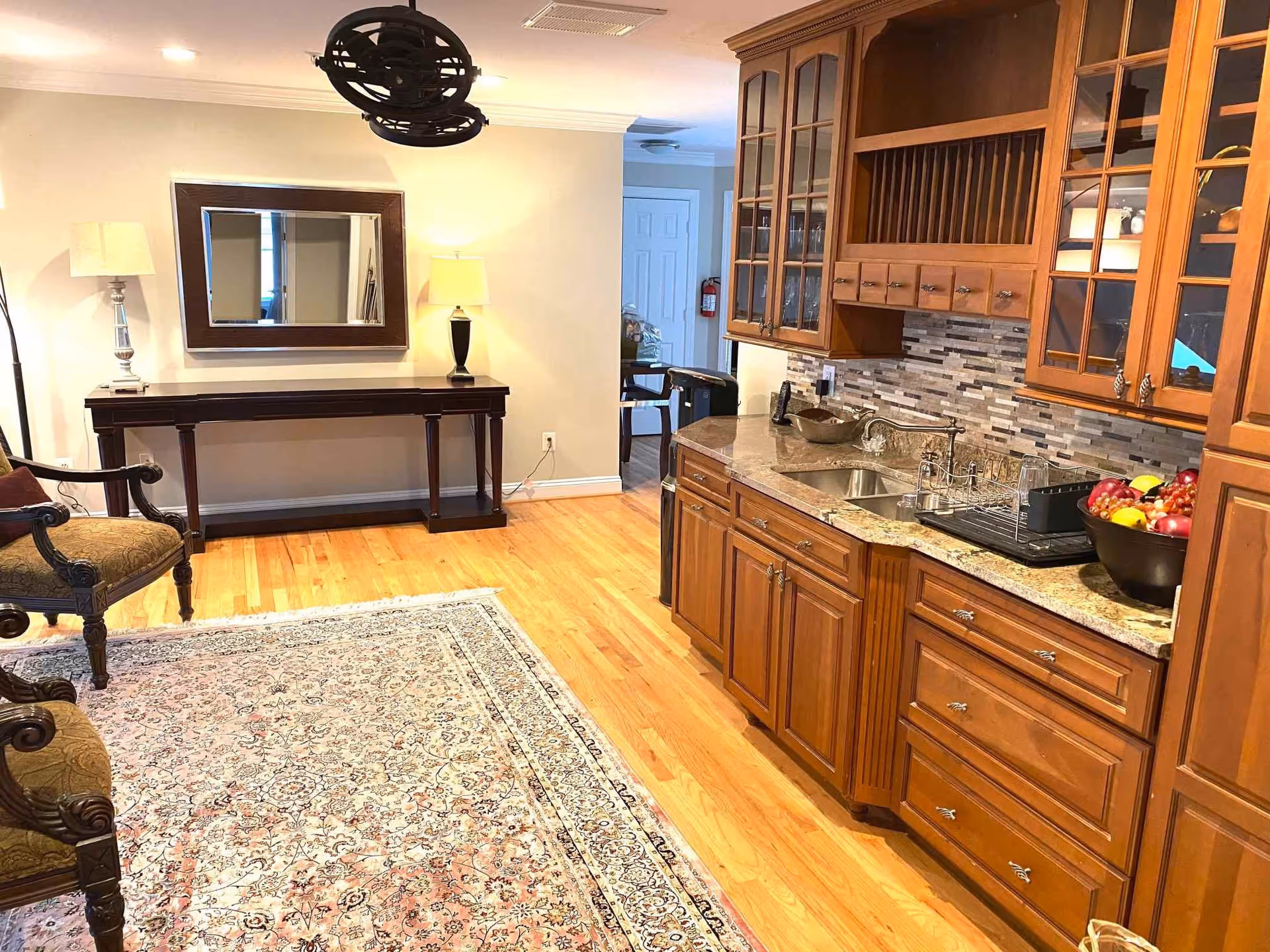 Open interior showing wooden kitchen cabinets and sink beside a seating area with rug, chairs, and a console table with lamps and mirror.