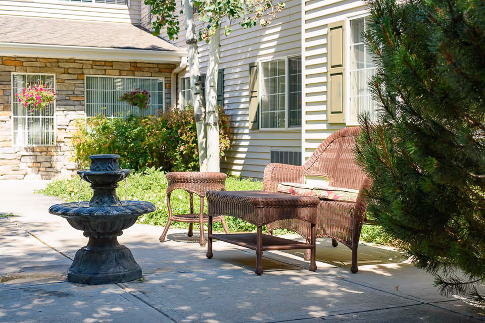 Outdoor patio area with wicker furniture including a loveseat, two chairs, and a coffee table. There is a small decorative water fountain on the concrete patio. The background shows the exterior of a building with stone and siding walls, windows with shutters, and hanging flower baskets.