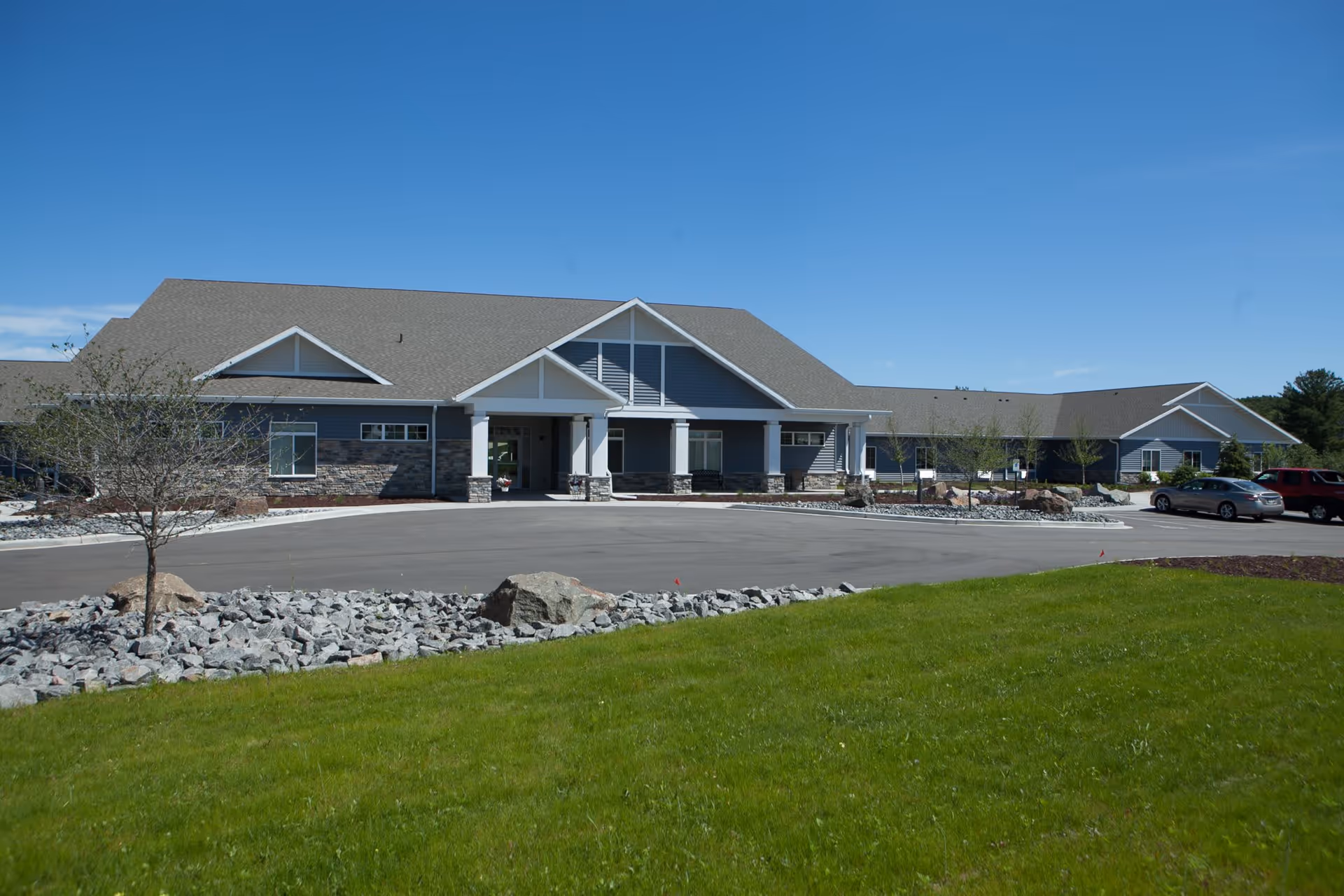Front exterior view of a single-story assisted living facility building with a gray roof, blue siding, stone accents, and a covered entrance. There is a paved driveway and parking area with a few cars, surrounded by green grass, small trees, and landscaping rocks under a clear blue sky.