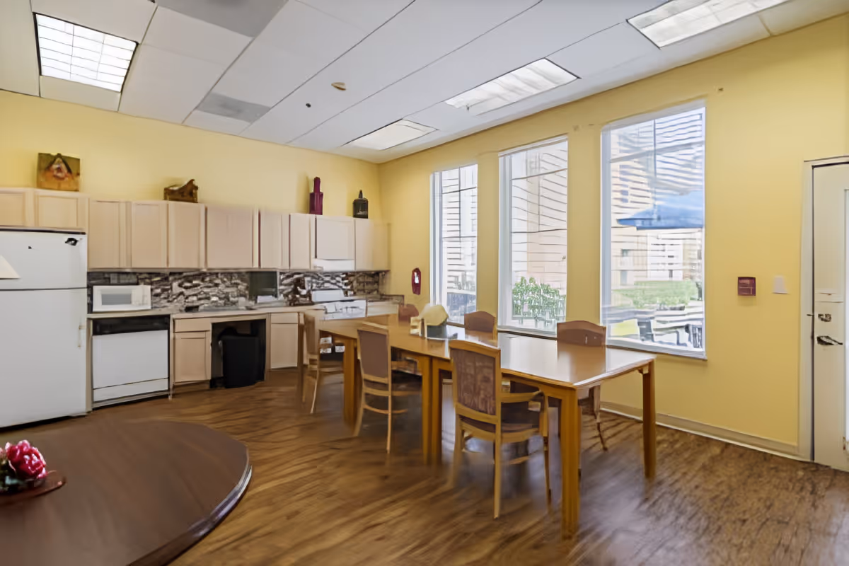 Community dining area with tables and chairs next to a small kitchenette and large windows.