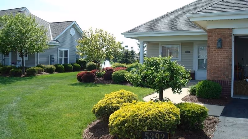 A well-maintained outdoor area of a senior living facility with green grass, trimmed bushes, small trees, and two single-story buildings with gray siding and shingled roofs under a clear sky.